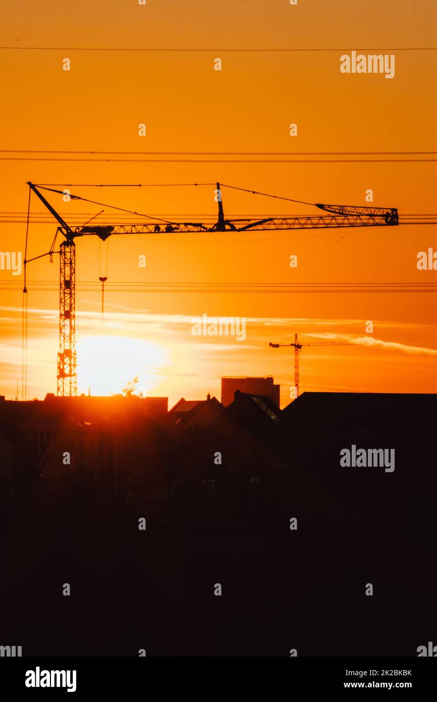 Tall construction crane silhouette in orange sky sunset shows construction site with engineering for modern buildings and city development as architectural teamwork for skyscrapers high voltage lines Stock Photo