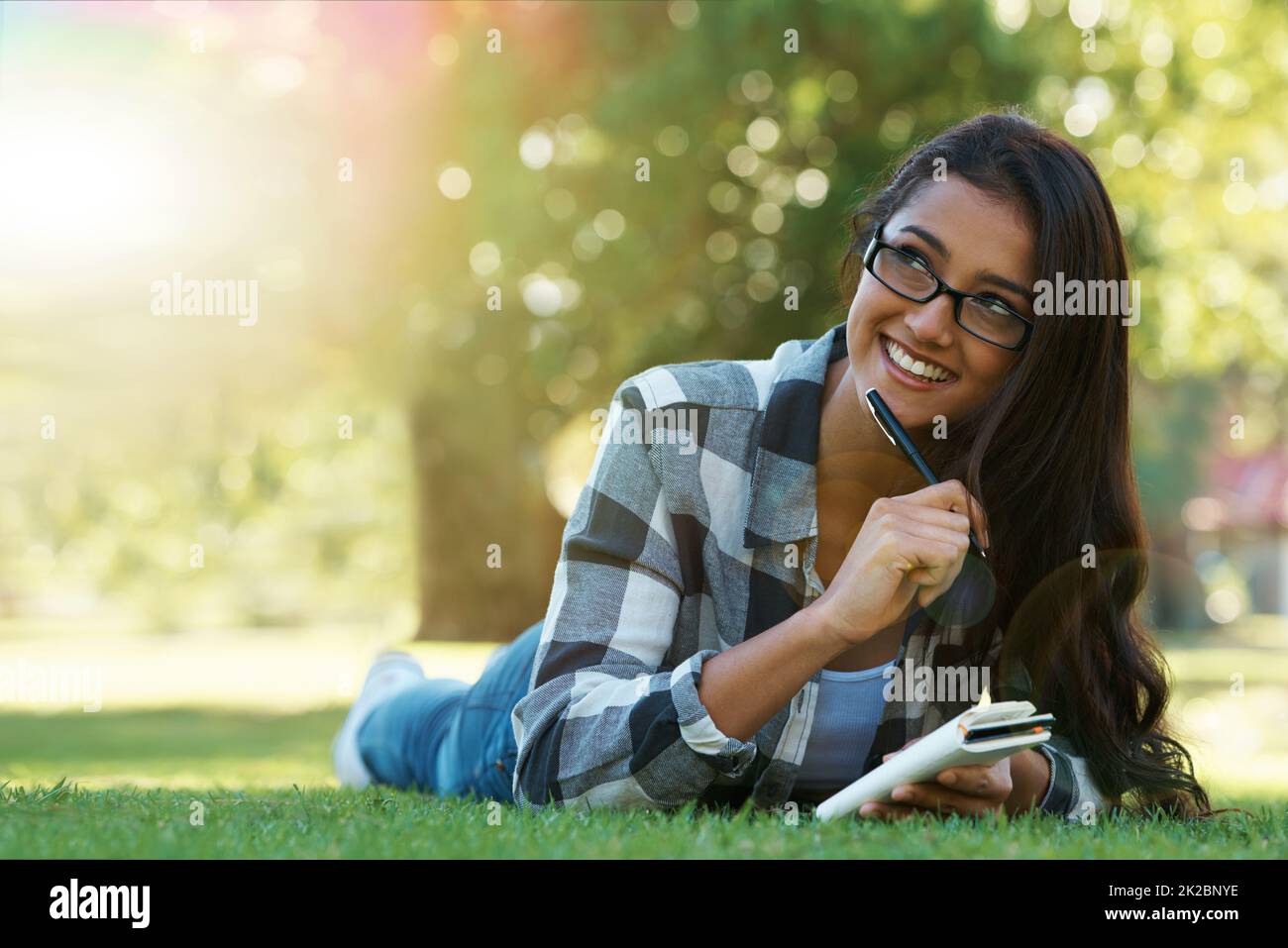 Jotting down her thoughts. A young woman lying on the grass writing in a notebook. Stock Photo
