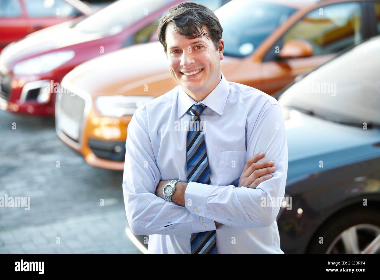 He can give you a great car deal. A man smiling confidently while standing in the lot of his dealership. Stock Photo