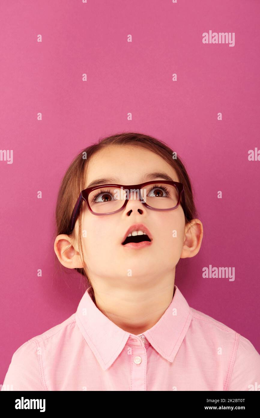 Childlike wonder. A little girl wearing spectacles looking up against a pink background. Stock Photo