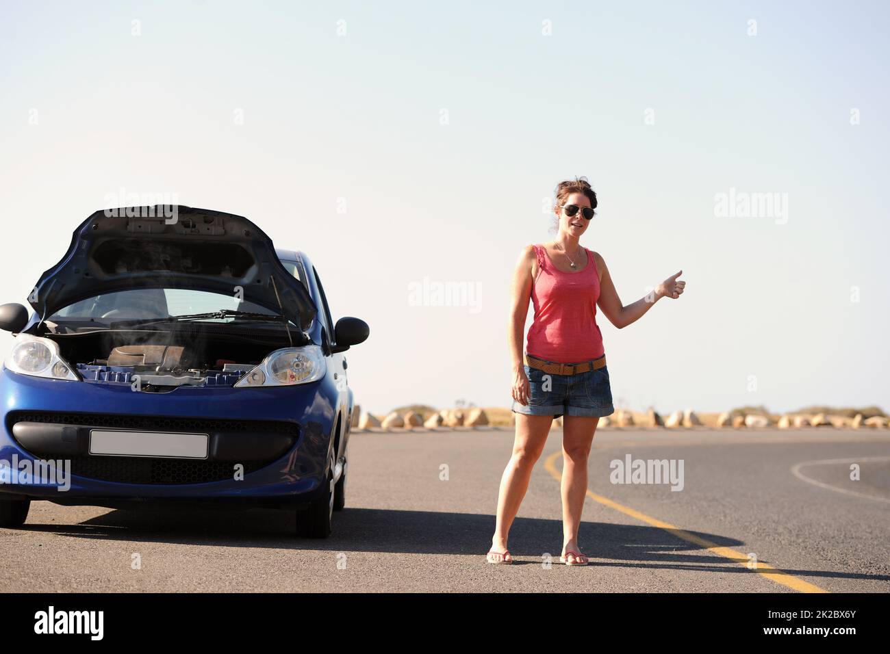 Shes in need of some road side assistance. A beautiful young woman standing with her broken-down car hitching a ride on the side of the road. Stock Photo