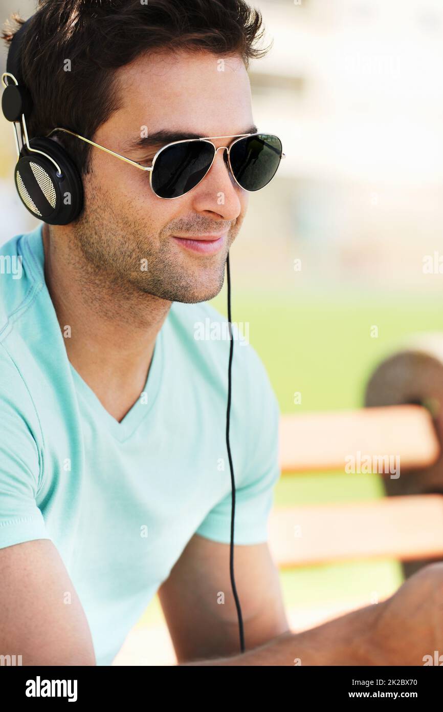 Tuning out to his favorite tunes. A smiling young man wearing sunglasses sitting on a bench in a park and listening to music through his headphones. Stock Photo