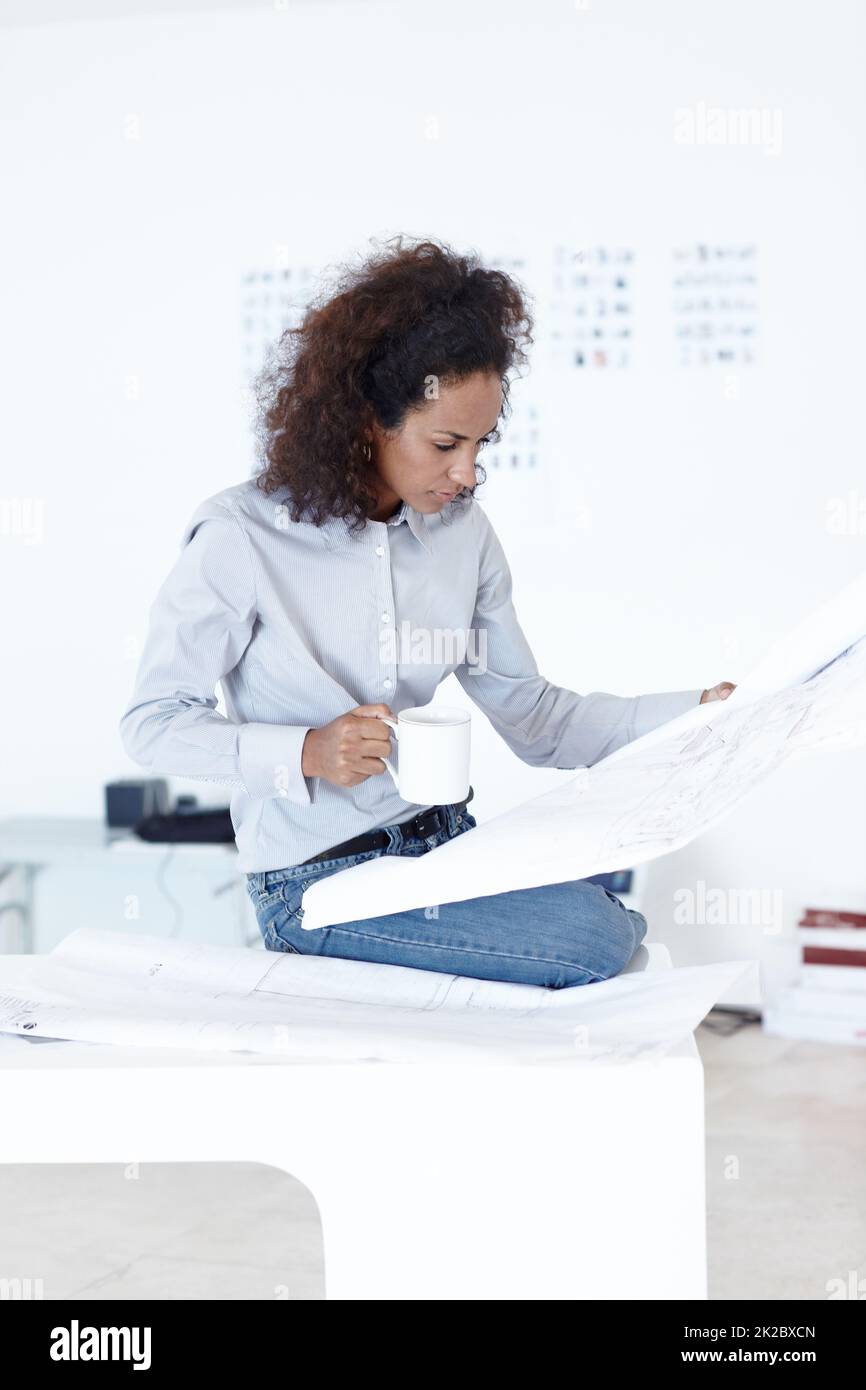Working on her latest project. Shot of a female architect drinking coffee while examining blueprints in her office. Stock Photo