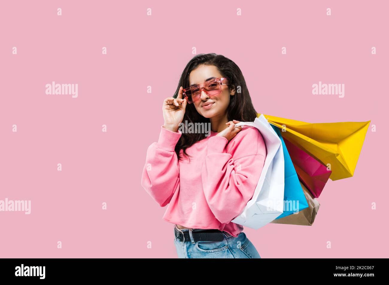 Shopping concept. Trendy pretty caucasian stylish brunette young woman, wearing casual clothes and pink sunglasses, holding shopping bags, stands on isolated pink background, looks at camera, smiling Stock Photo