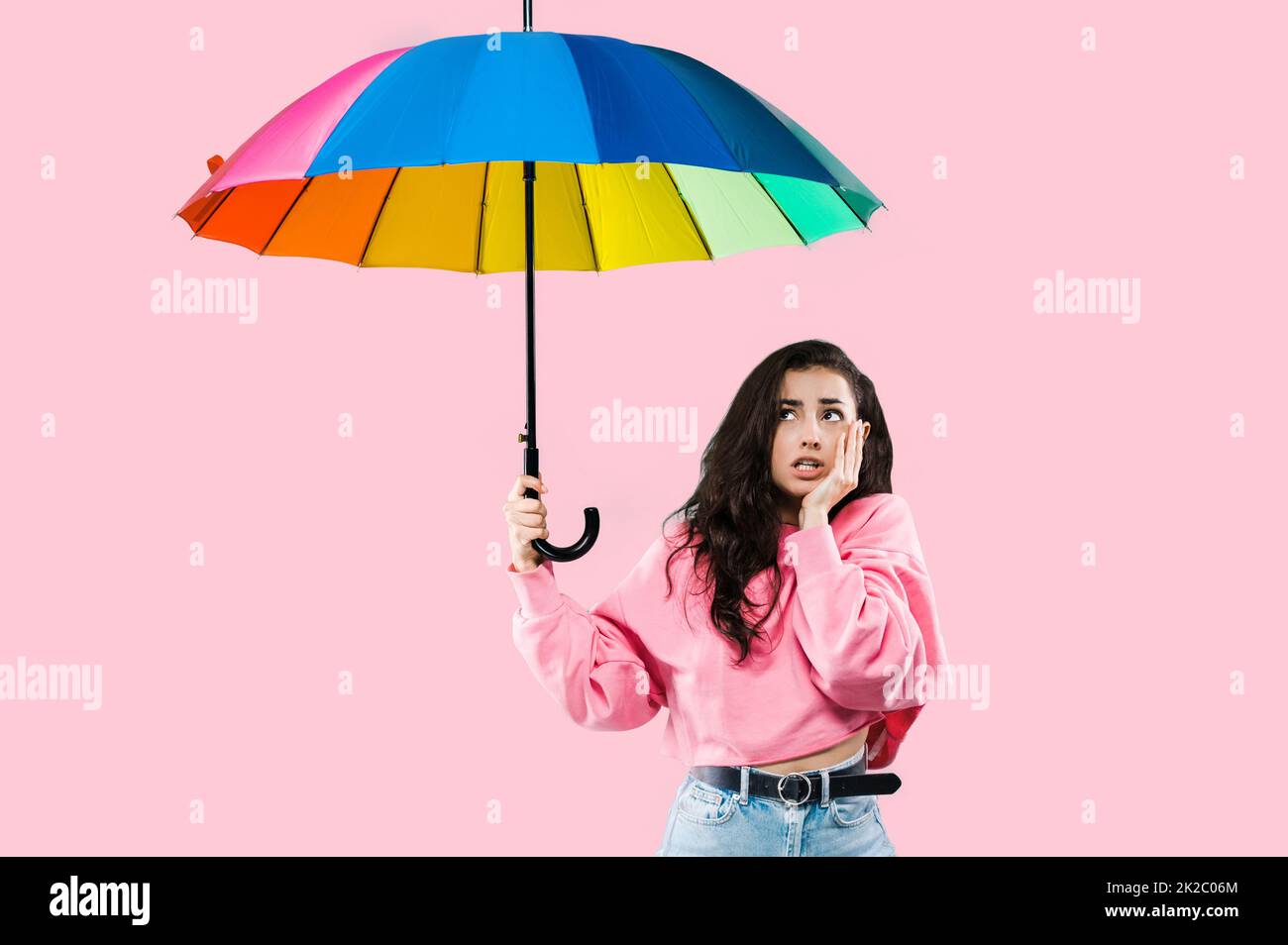 Worried caucasian upset young woman, stylishly dressed, holding an open rainbow umbrella in her hand, looking up, waiting for rain, standing over isolated pink background Stock Photo