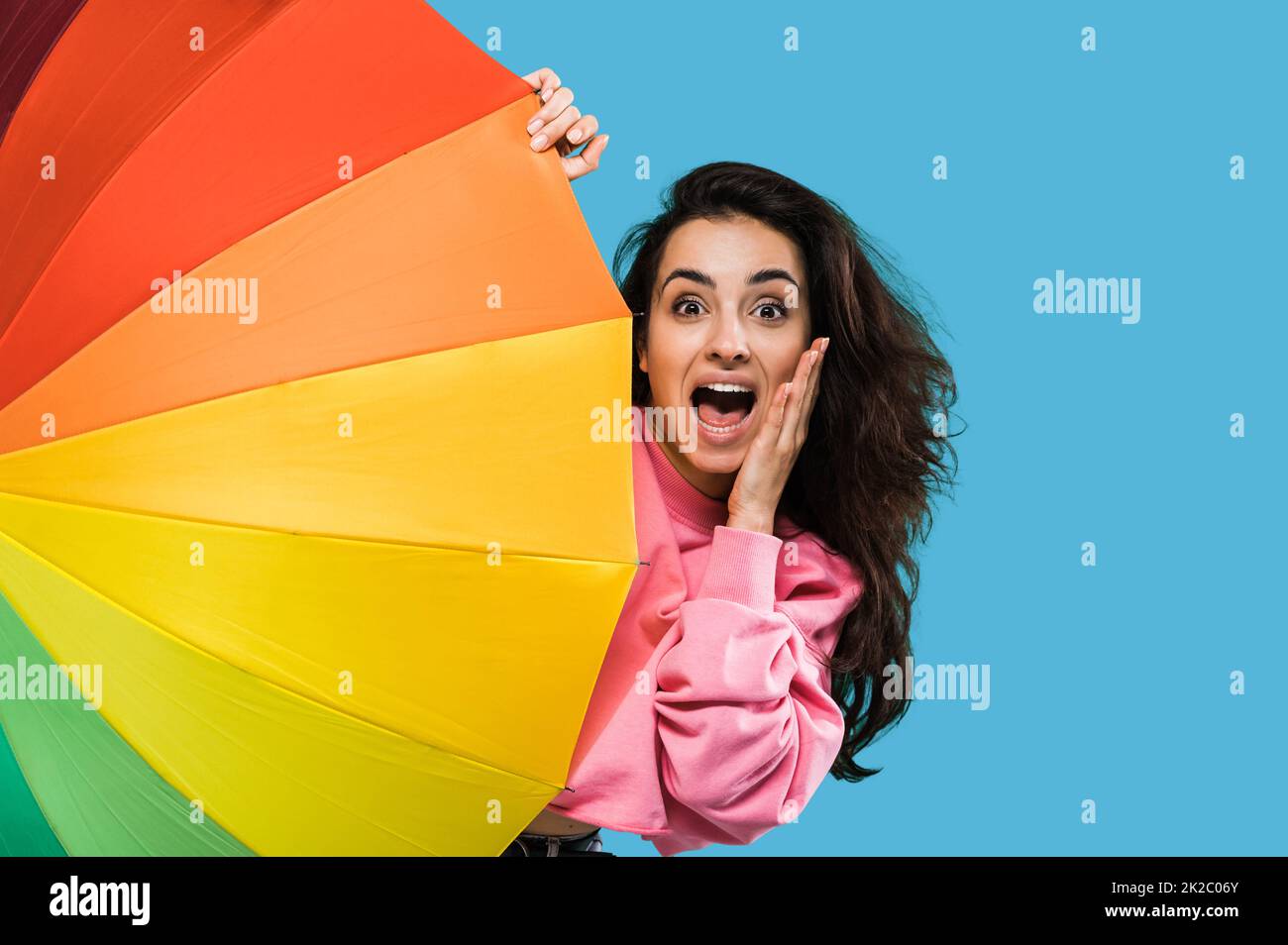 Shocked excited caucasian beautiful long haired young woman in pink sweater, peeking out from behind colorful umbrella, looking at camera in amazement, standing over isolated blue background Stock Photo