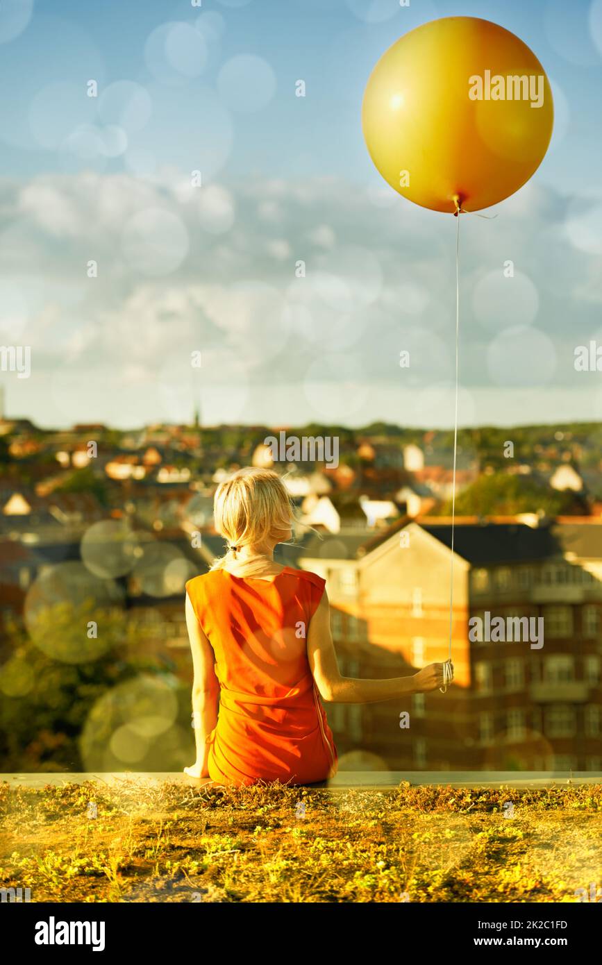 Reliving her childhood. Rear view of a young woman sitting with a balloon and a city in the background. Stock Photo
