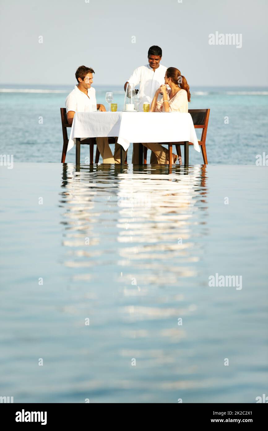 Surrounded by the beauty of nature. A young couple sitting at a table outside surrounded by water and being served wine. Stock Photo