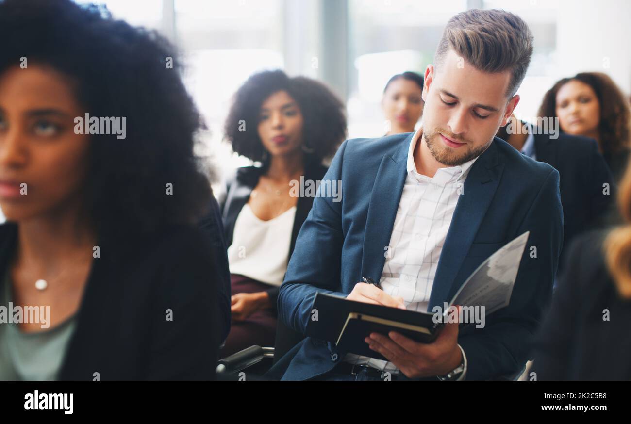 Lots to take in, you may want to take note. Shot of a young businessman taking down notes while sitting in the audience of a business conference. Stock Photo
