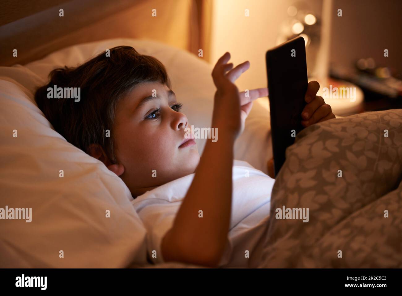 Some light reading before bed. Shot of a young boy playing on a tablet while lying in bed. Stock Photo