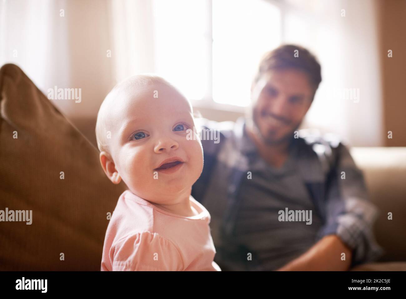 Already too cute and curious. Shot of an adorable baby girl sitting on the sofa with her father in the background. Stock Photo