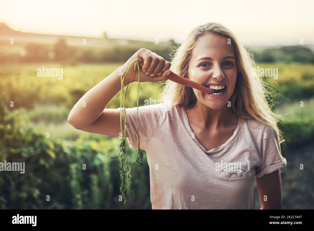 Delicious and nutritious. Cropped portrait of a young woman holding up a carrot and taking a bite with her farmland in the background. Stock Photo