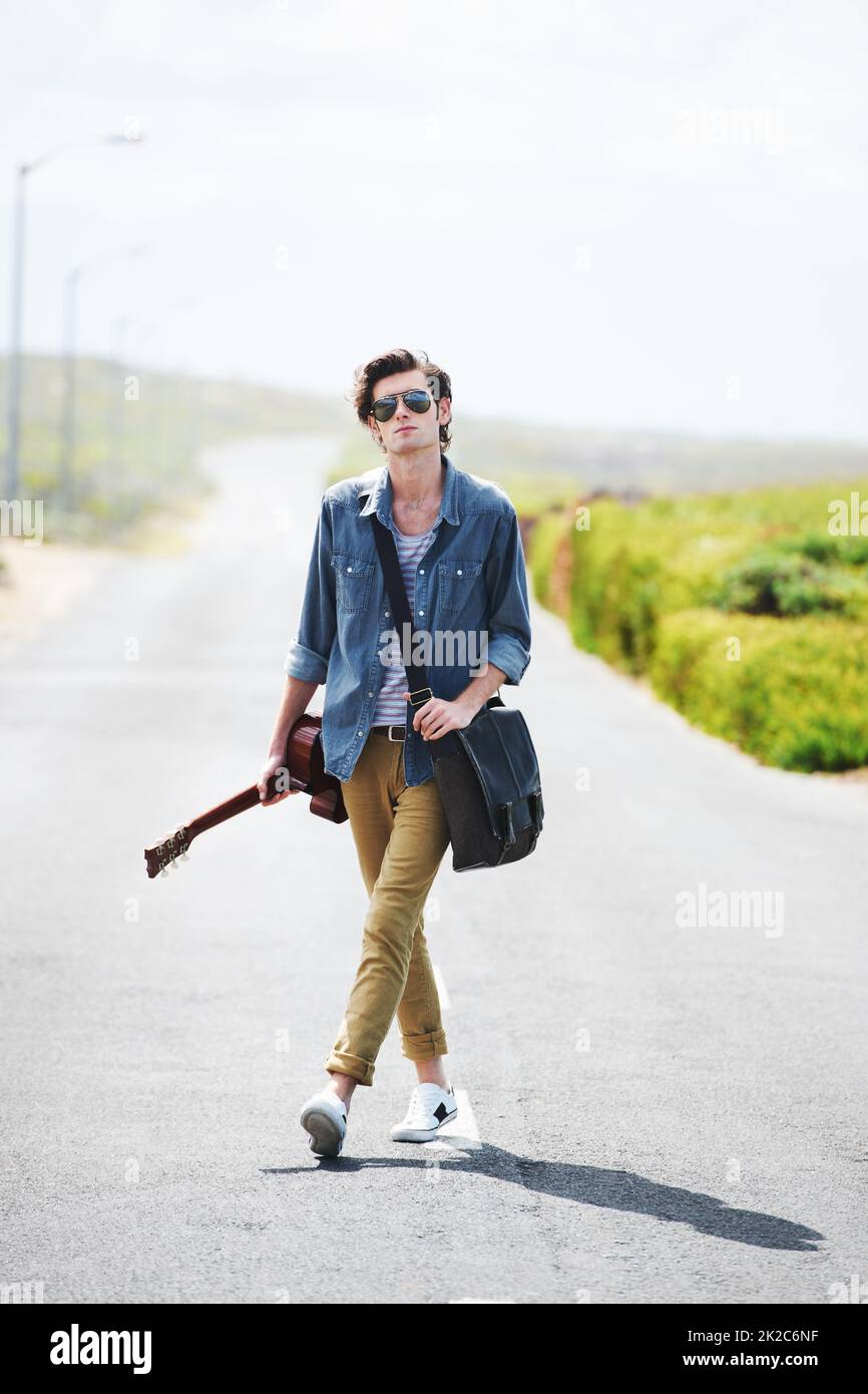 All I need is my guitar and the open road. Trendy young male wearing sunglasses standing in the middle of the road holding a guitar. Stock Photo