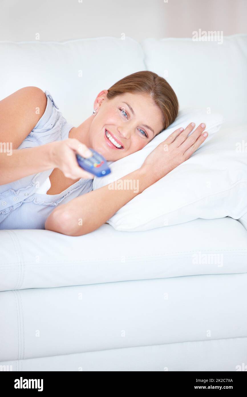 Channel-hopping is so relaxing. Smiling young woman relaxing at home on the couch using her TV remote - portrait. Stock Photo