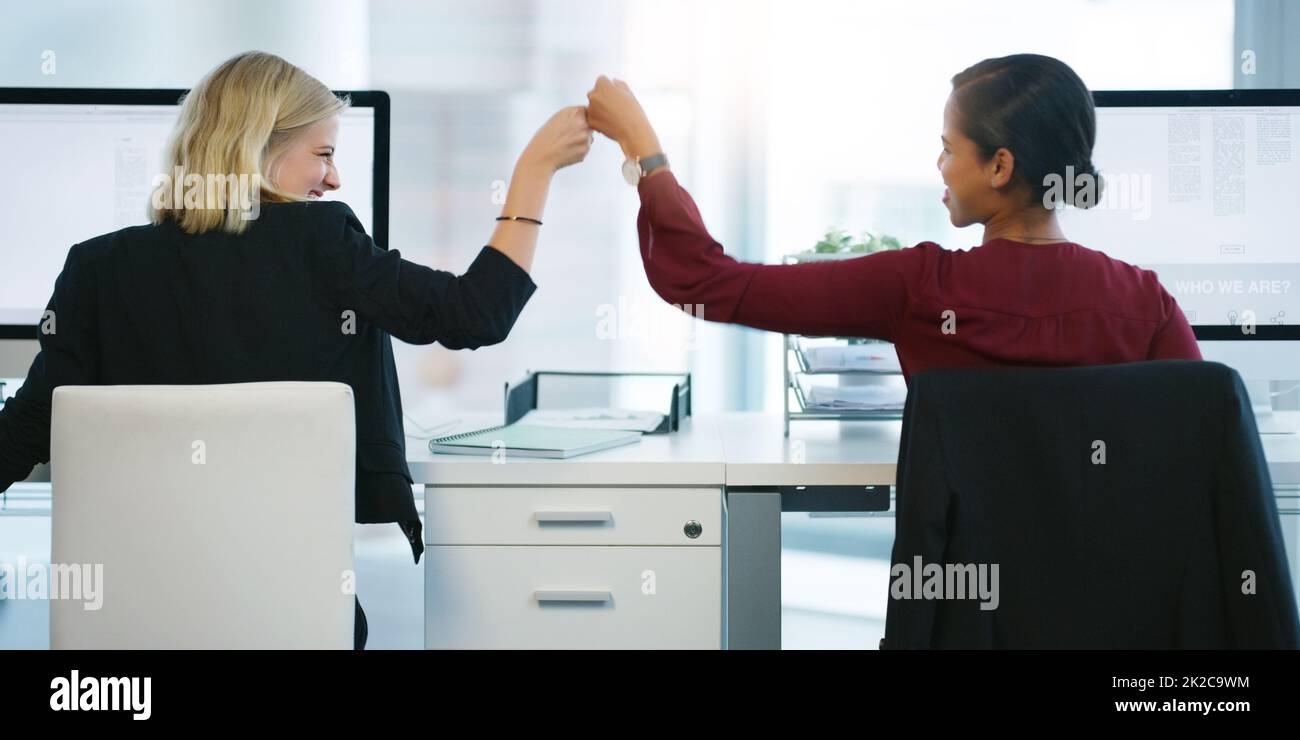 Today we outdid ourselves. Rearview shot of two young businesswomen pounding their fists in succession while being seated at their desk in the office. Stock Photo