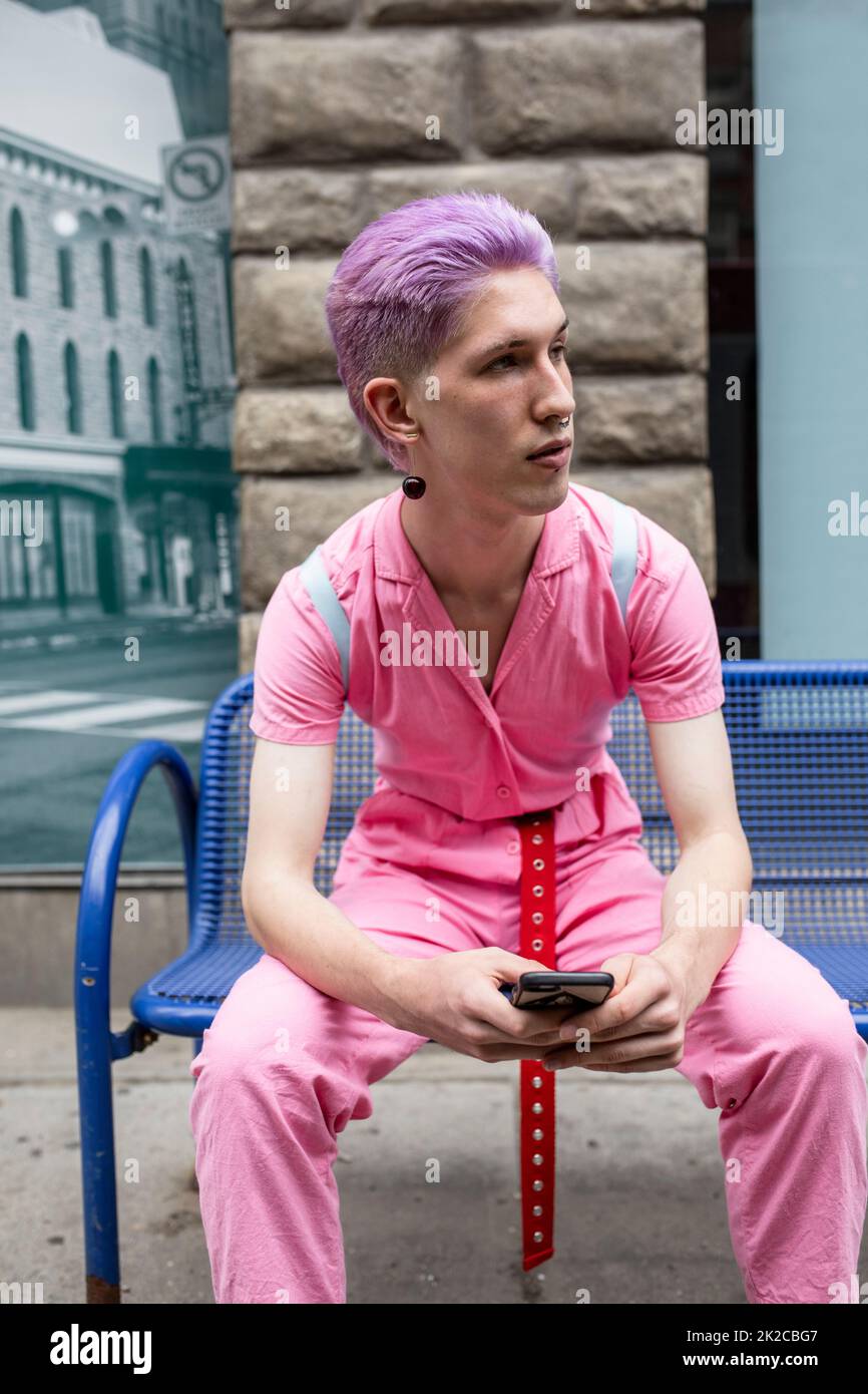 Stylish nonbinary young adult in pink jumpsuit on city bench Stock
