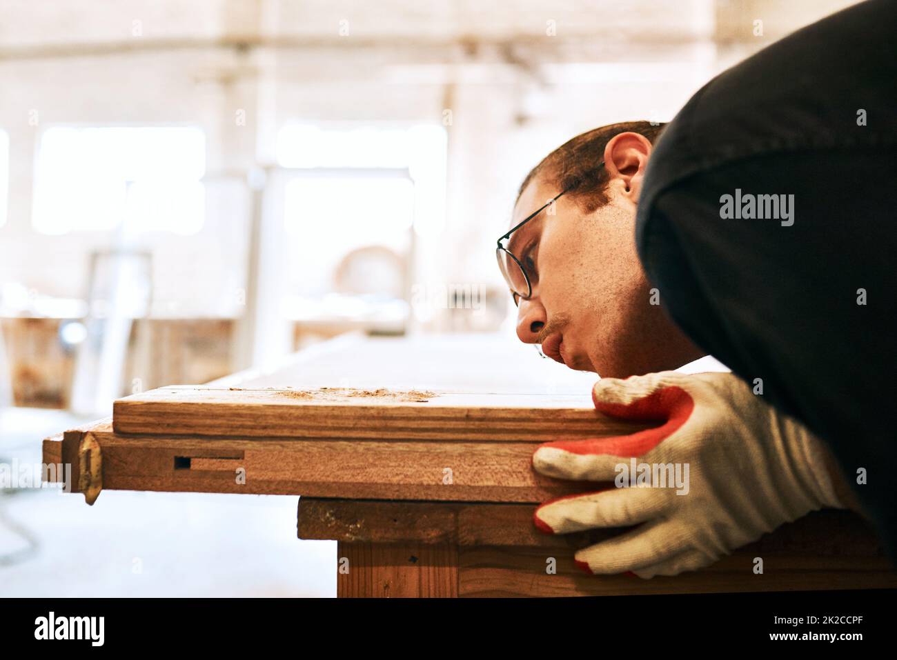 I like having a dust-free working environment. Shot of a handsome young carpenter blowing dust off a wooden plank inside a workshop. Stock Photo