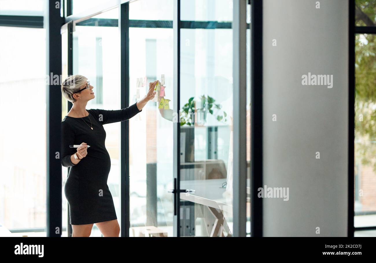 This plan will surely work out well. Shot of a pregnant businesswoman brainstorming with notes on a glass wall in an office. Stock Photo