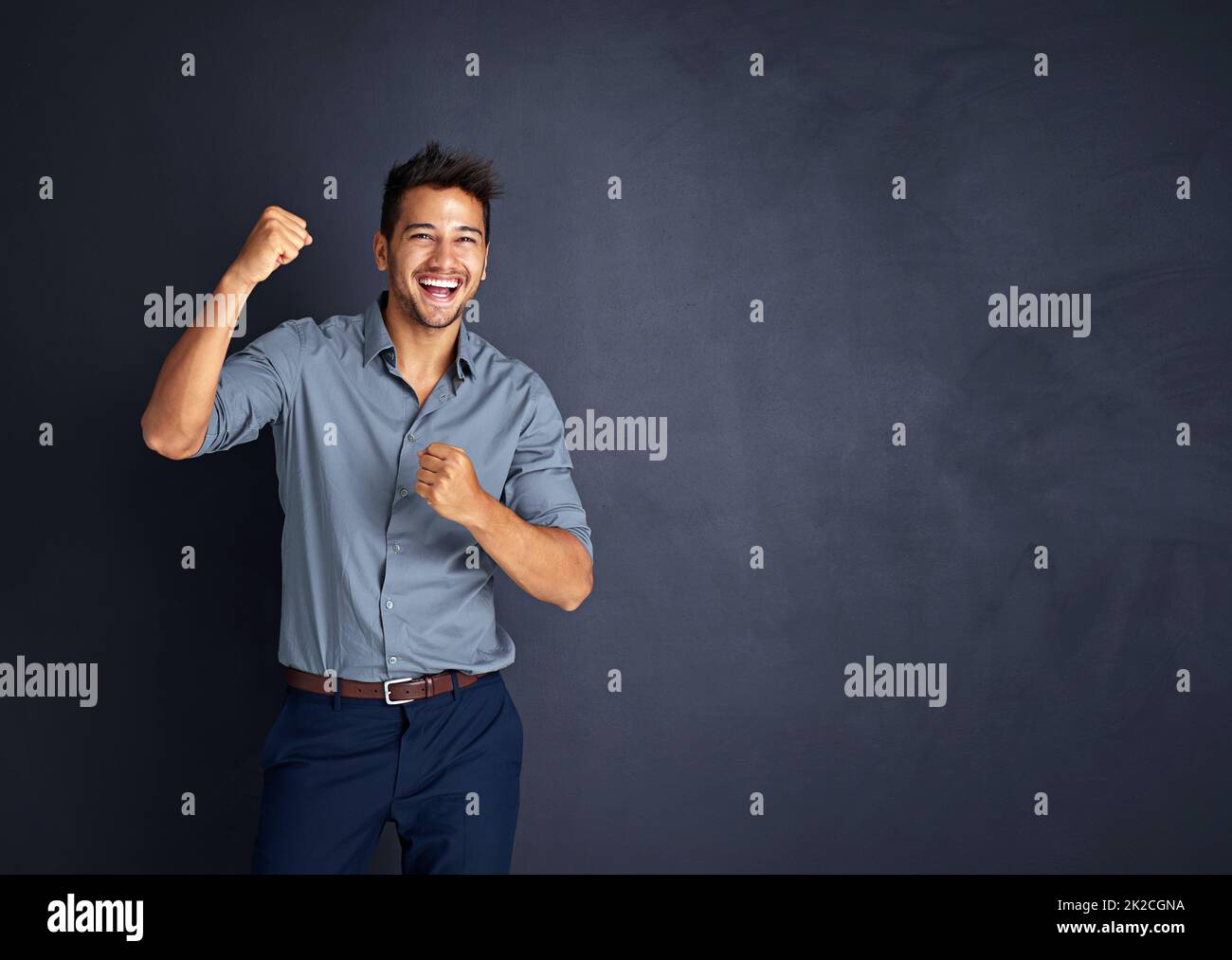 Cheering on through all his achievements. Studio shot of a young businessman standing with his fists raised in celebration against a dark background. Stock Photo