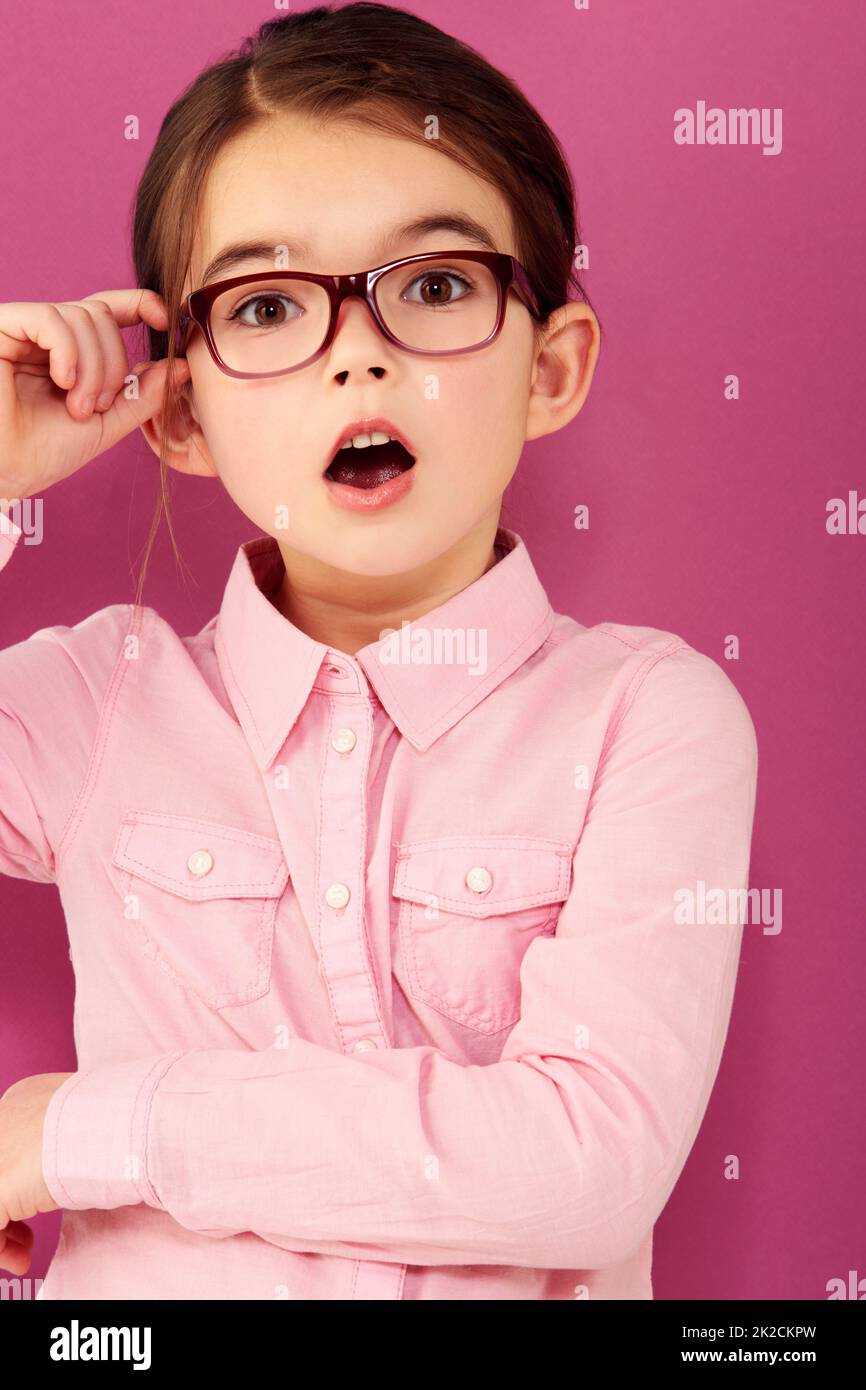 Shes seriously surprised. A little girl with expression of surprise holding the edge of her spectacles against a pink background. Stock Photo