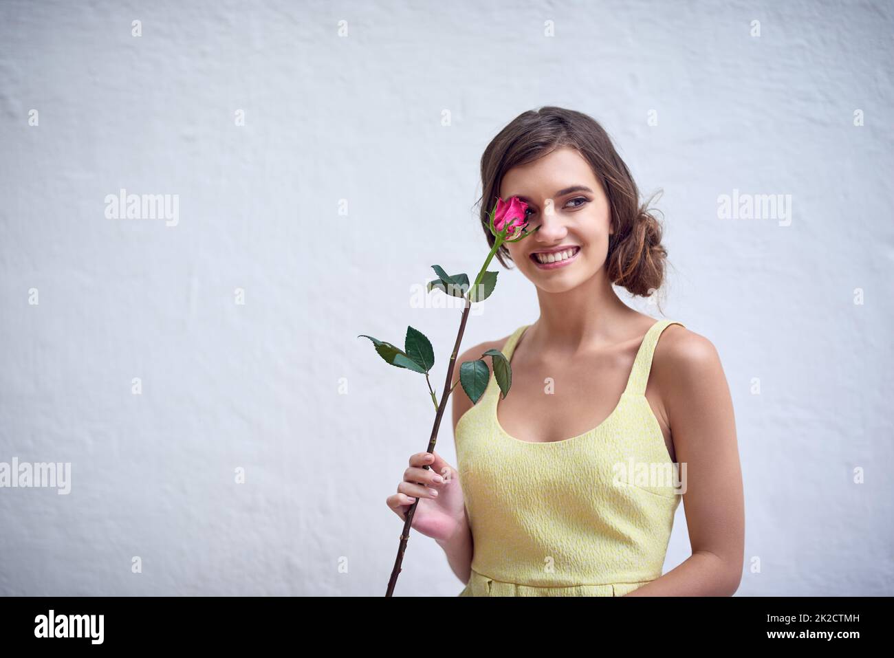 Shes a flower type of girl. Studio shot of a cheerful young woman holding a pink rose next to her face while standing against a grey background. Stock Photo