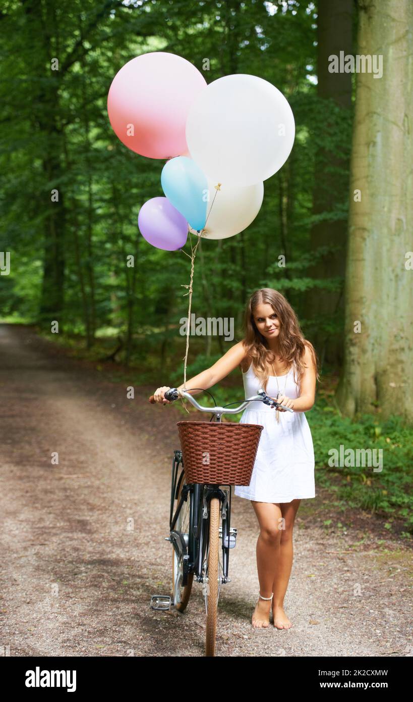 Feeling a little whimsical. A pretty young woman with a bunch of balloons and a bicycle walking in the forest. Stock Photo