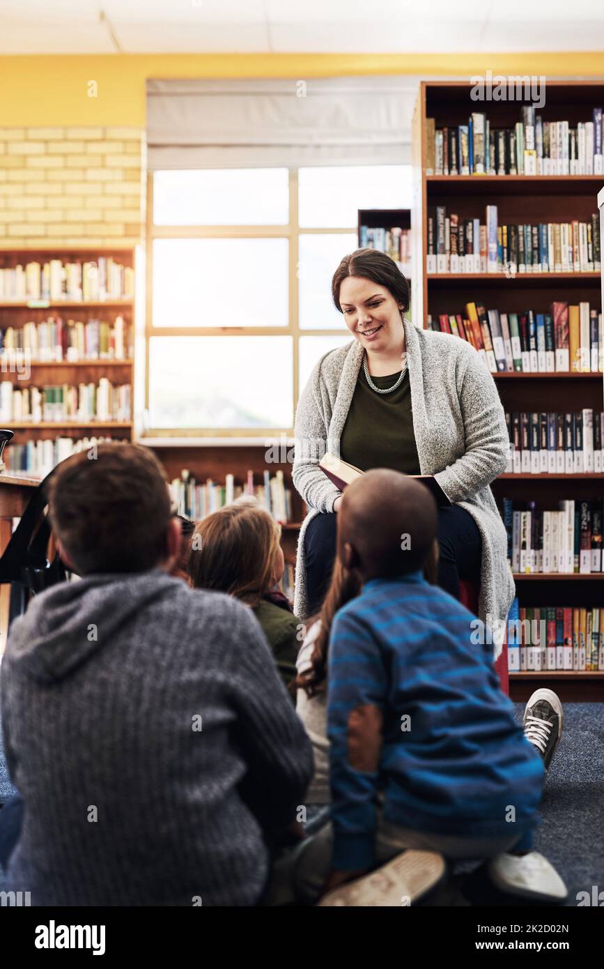 Shes sharing her love for books to the class. Shot of a teacher reading to a group of elementary school kids in the library. Stock Photo