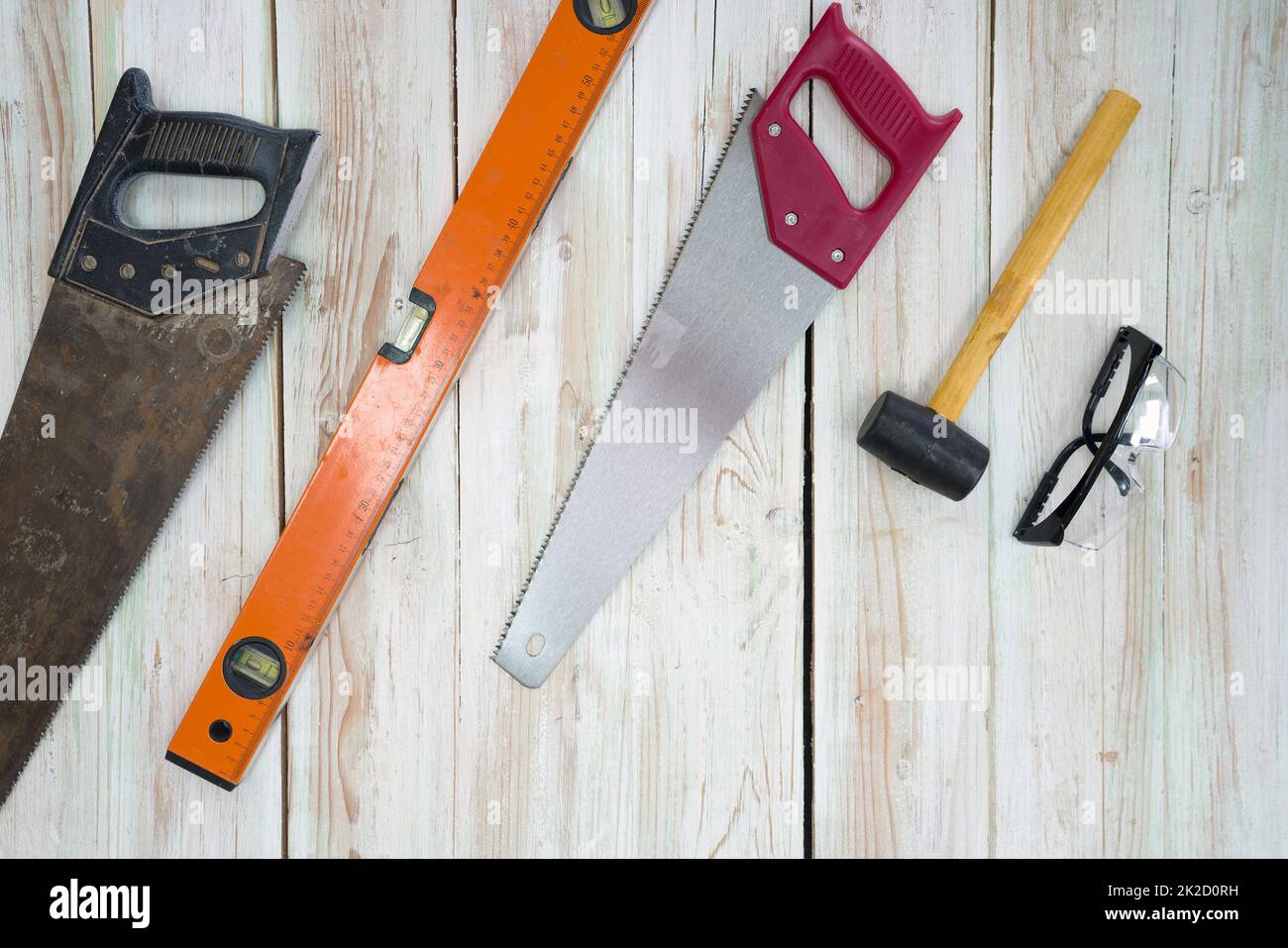 The equipment for woodworking is organized neatly on the wood floor. Ordered as follows, old saw full of rust, orange magnetic aluminum level, red hand saw, yellow mallet, black dustproof glasses. Stock Photo