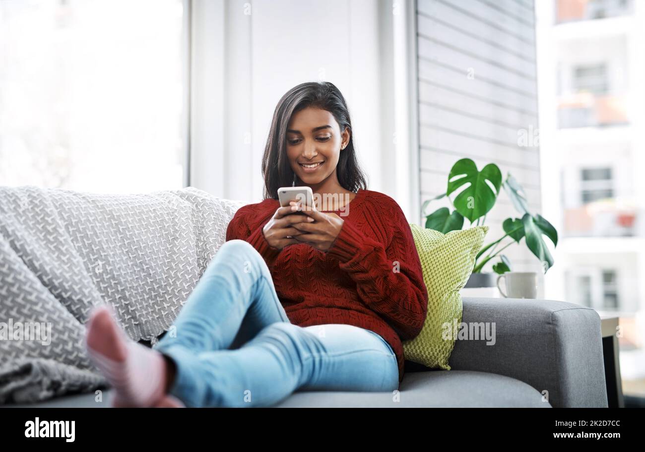 Catching up on social media this weekend. Cropped shot of a beautiful young woman using a cellphone while chilling on the sofa in the living room at home. Stock Photo