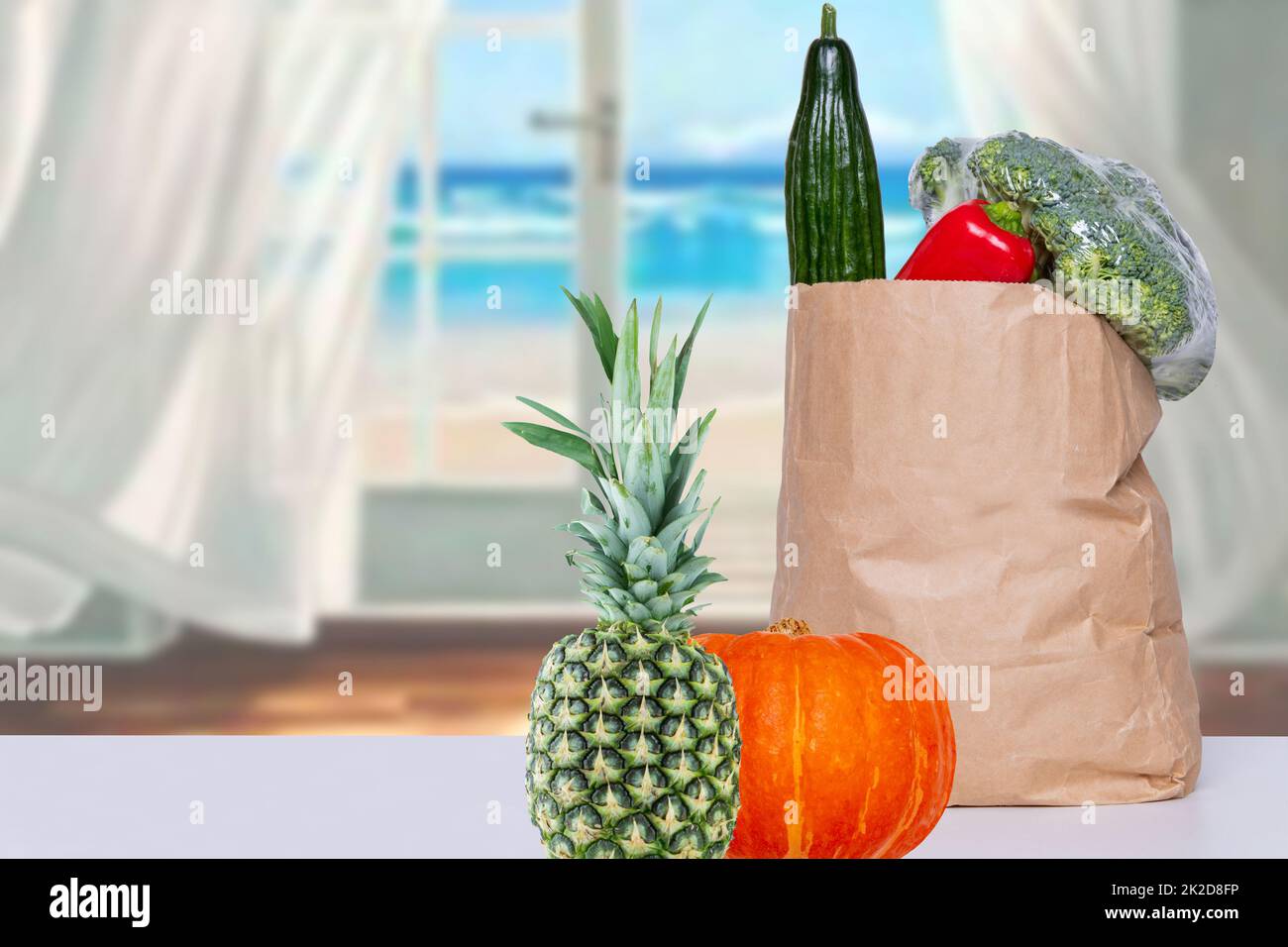 Close up of a brown paper shopping bag with fresh vegetables, a pumpkin and a pineapple on the table over an abstract blurred sea background. Concept of strengthening the immune system. Copy space. Stock Photo