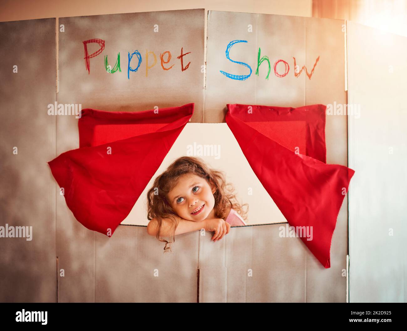 Fostering a childs imagination through play. Shot of an adorable little girl standing in the window of a puppet show theatre at home. Stock Photo