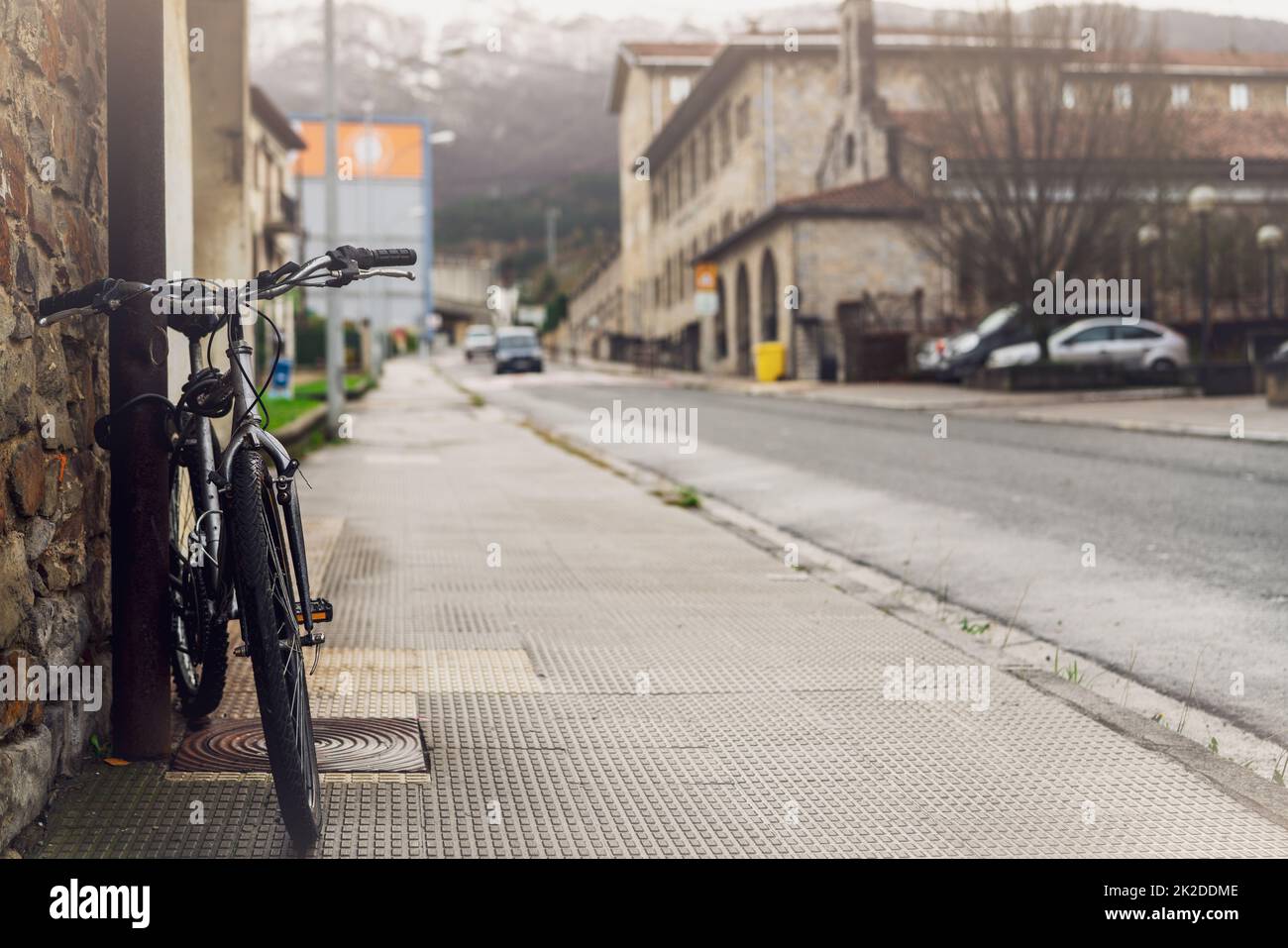 Bicycle parked on sidewalk near city street in Spain. Bike lean on pole beside old building. Front view of bicycle on blurred building, car driving on the road, and mountain background. Europe travel. Stock Photo