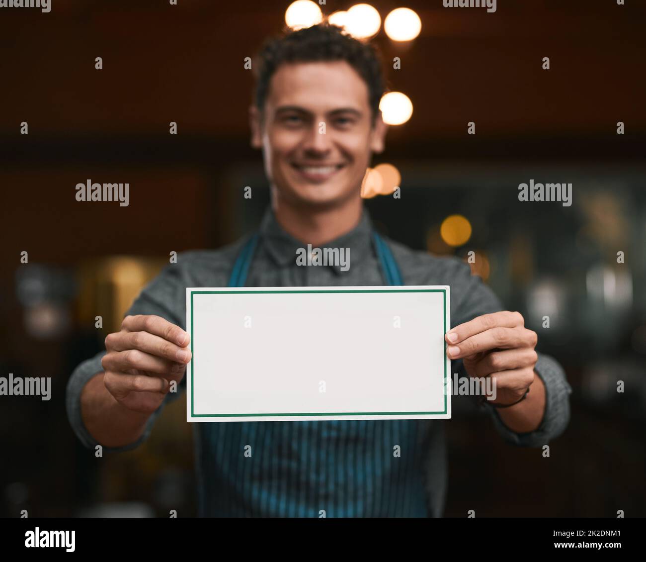 We will be glad if you can join us. Portrait of a cheerful young man holding a sign while standing inside of a beer brewery during the day. Stock Photo