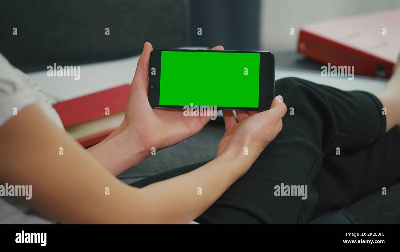 Young Woman sitting on sofa while looking at movies using phone in horizontal mode with mock up green screen chroma key display. Stock Photo