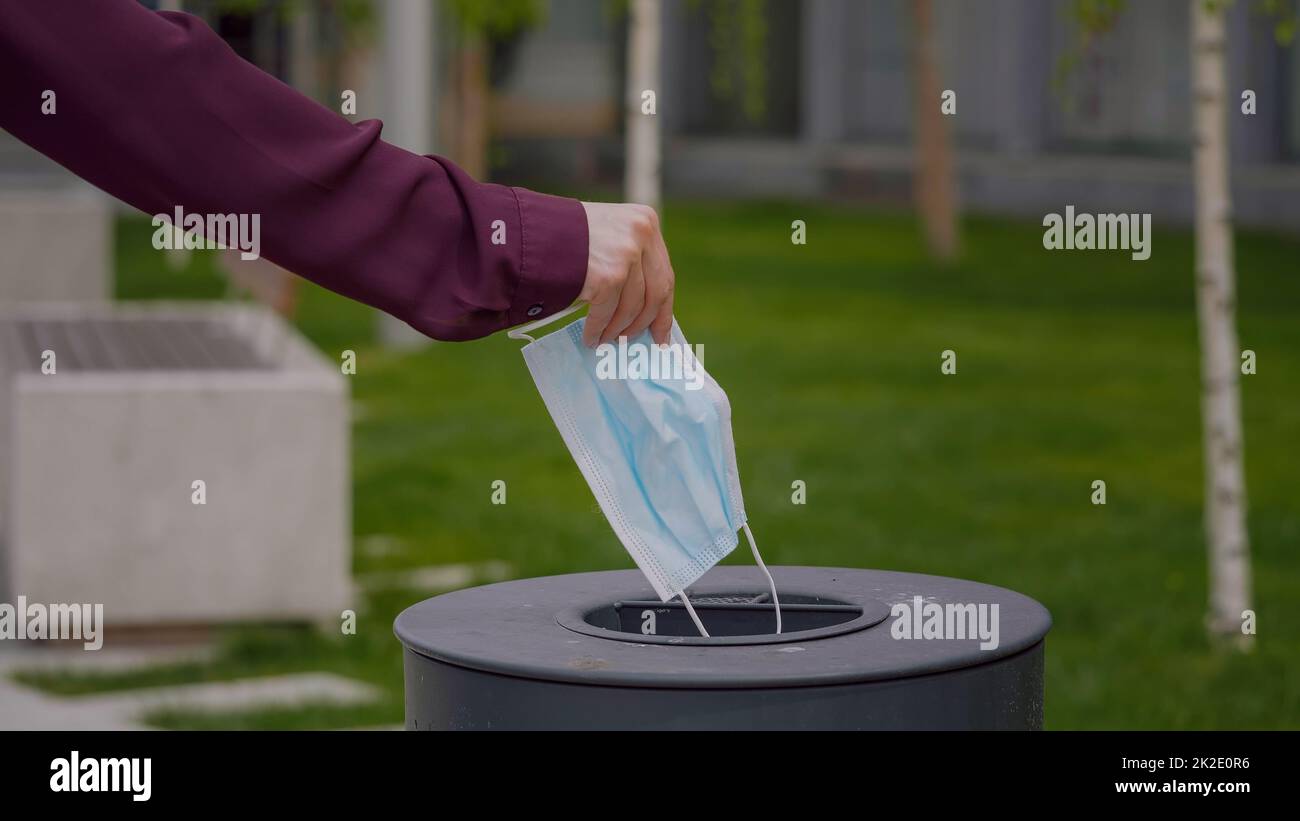 Woman throws a medical mask into the trash outdoors. The used face mask is dumped in a trash. Medical protective mask using to health protection from corona virus. Stock Photo