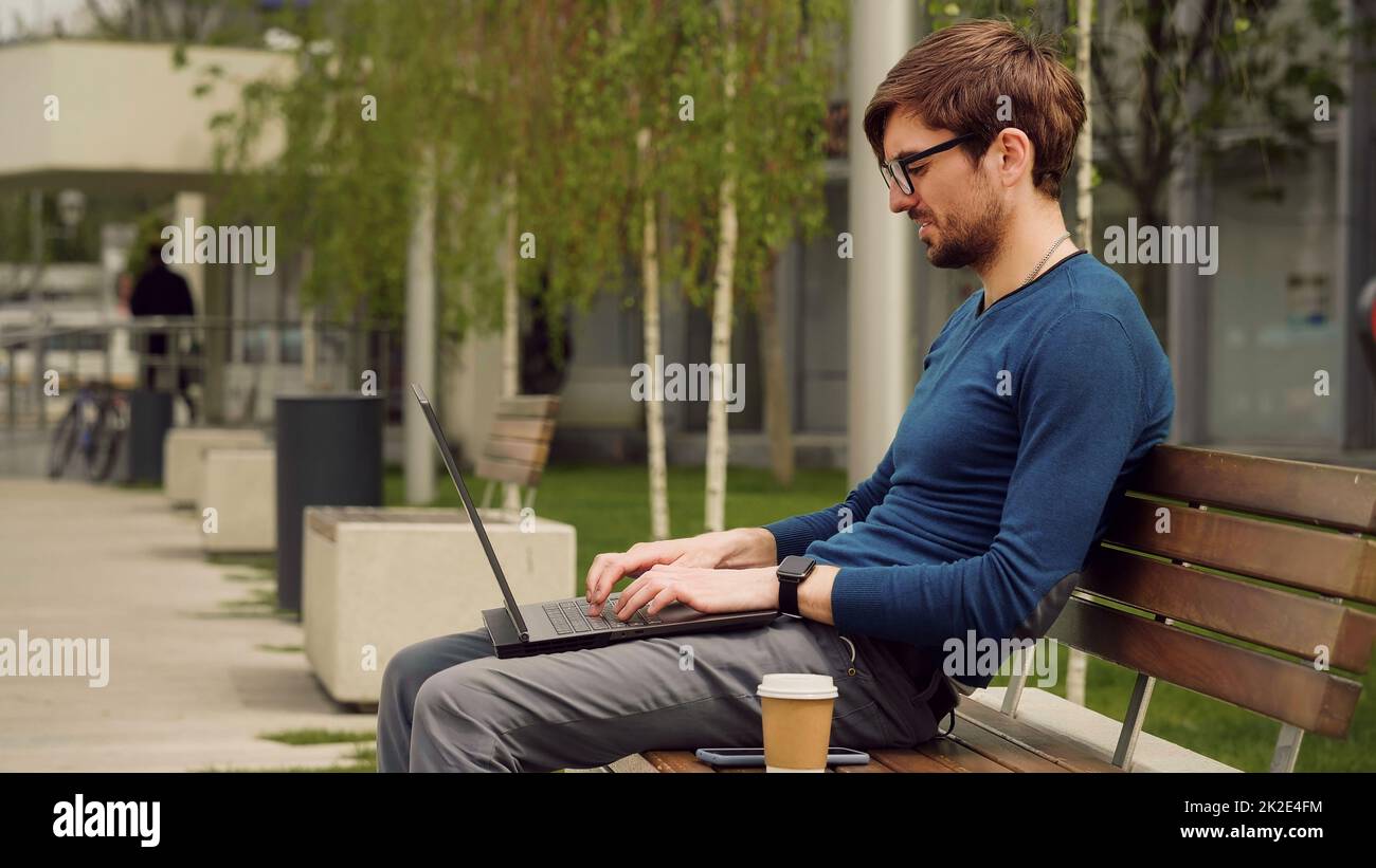 Young business man using laptop while sitting outdoor city park. Student studying on a bench. Entrepreneur working on laptop. Stock Photo