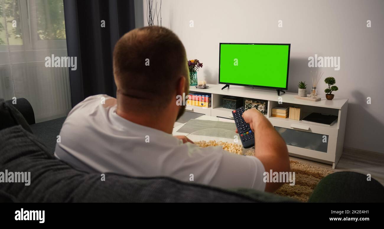 Man Watching Green Chroma Key Tv Screen and Changing Channels with Remote Control several times. Male sitting on the couch in the living room in the evening Stock Photo