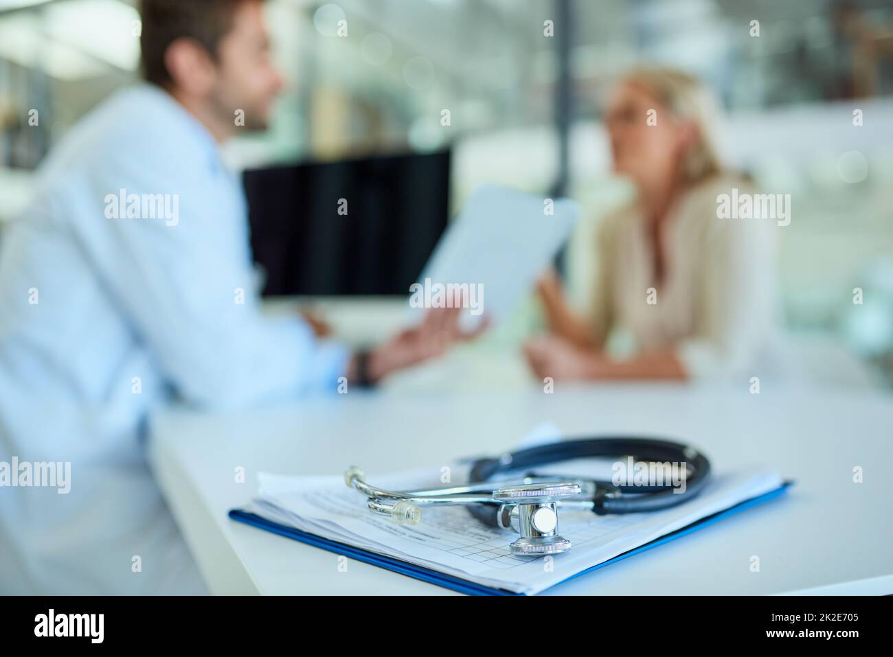 Were always here to listen to your health concerns. Shot of a stethoscope and a clipboard on a desk with a doctor and patient in the background. Stock Photo