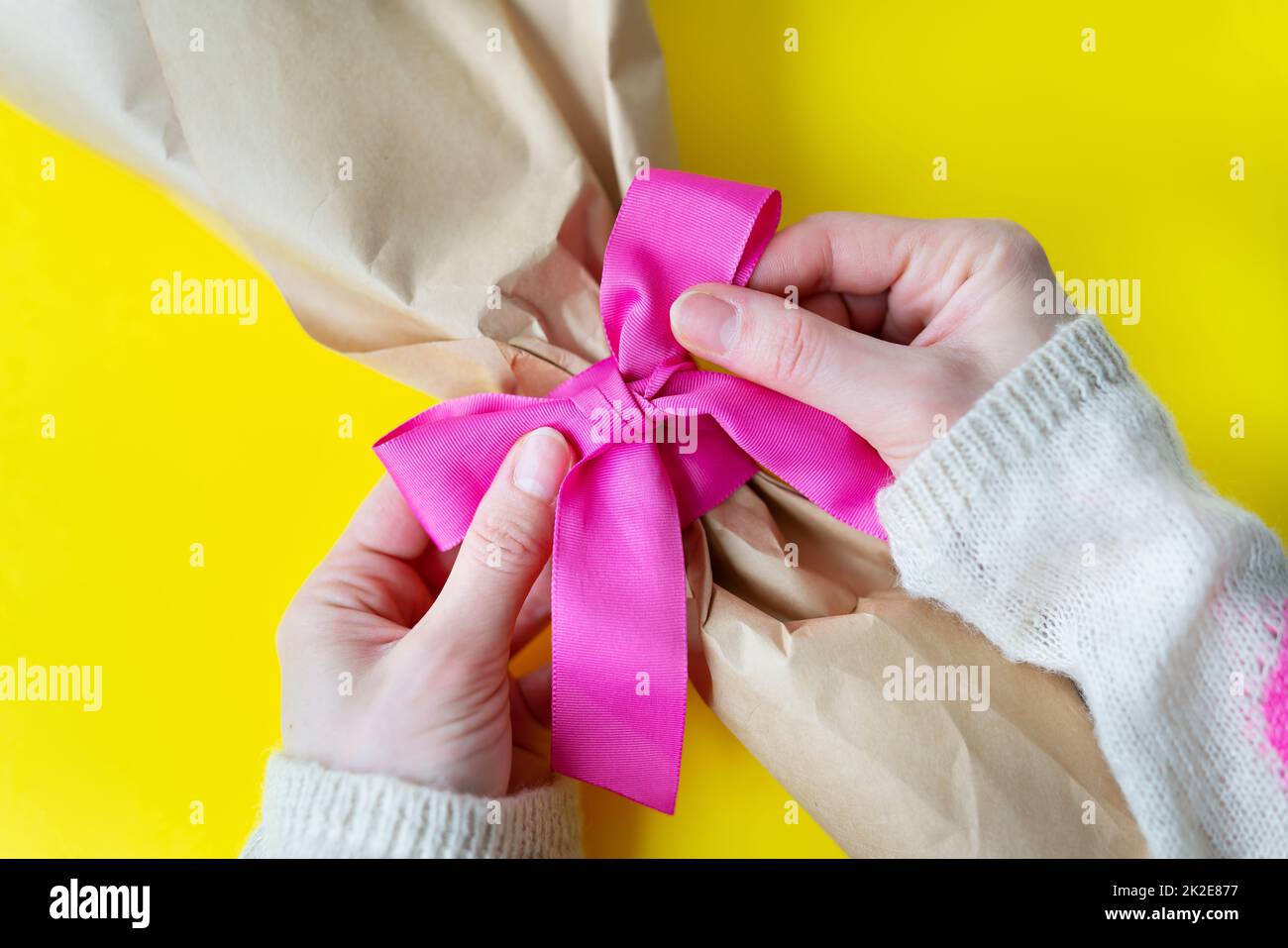 Top view of female hands tying a bouquet with a pink ribbon with gifts on a yellow background. Close-up, space for an inscription. Stock Photo