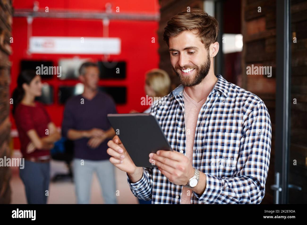 Enjoying his new work tool. Shot of a young designer using a tablet with her colleagues standing in the background. Stock Photo