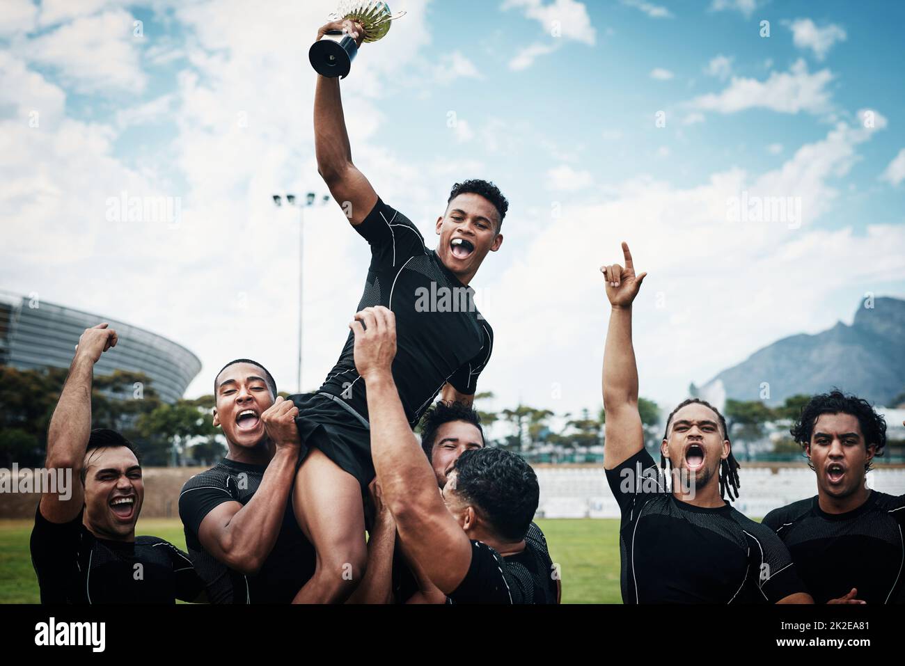 Theyre not just any team theyre rugby cup winners. Cropped shot of a handsome young rugby player holding up a trophy while celebrating with his team on the field. Stock Photo