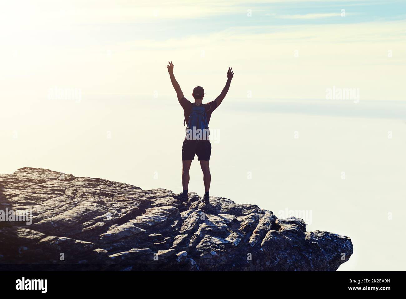 The best view comes after the hardest climb. Rearview shot of an unidentifiable young man raising his arms in triumph on a mountaintop. Stock Photo