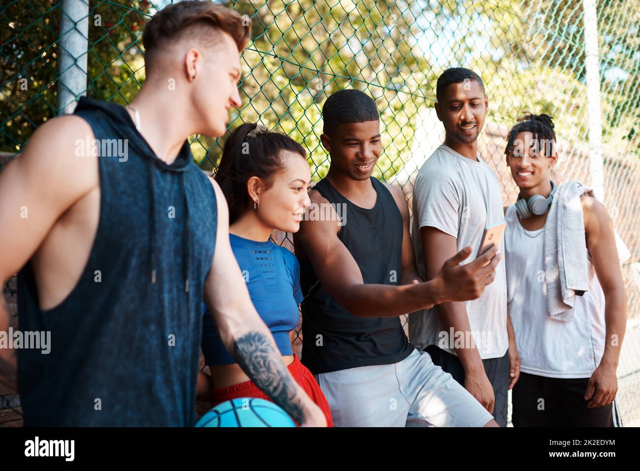 You guys have got to see this. Shot of a group of sporty young people looking at something on a cellphone while standing along a fence outdoors. Stock Photo