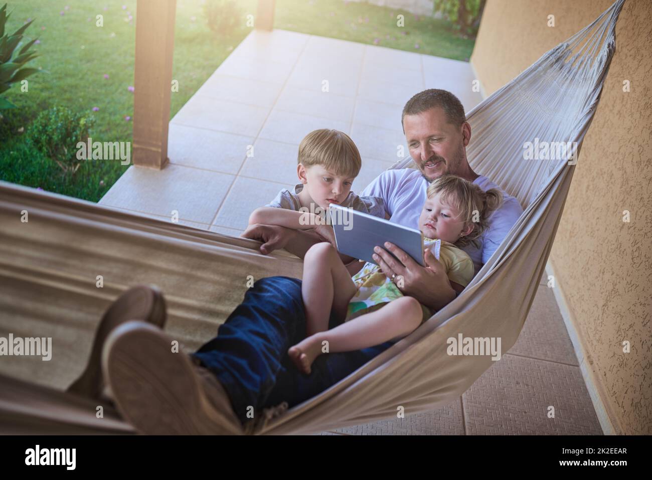 Swinging along to a chilled weekend with the kids. Shot of a father and his two little children using a digital tablet while relaxing on a hammock outdoors. Stock Photo