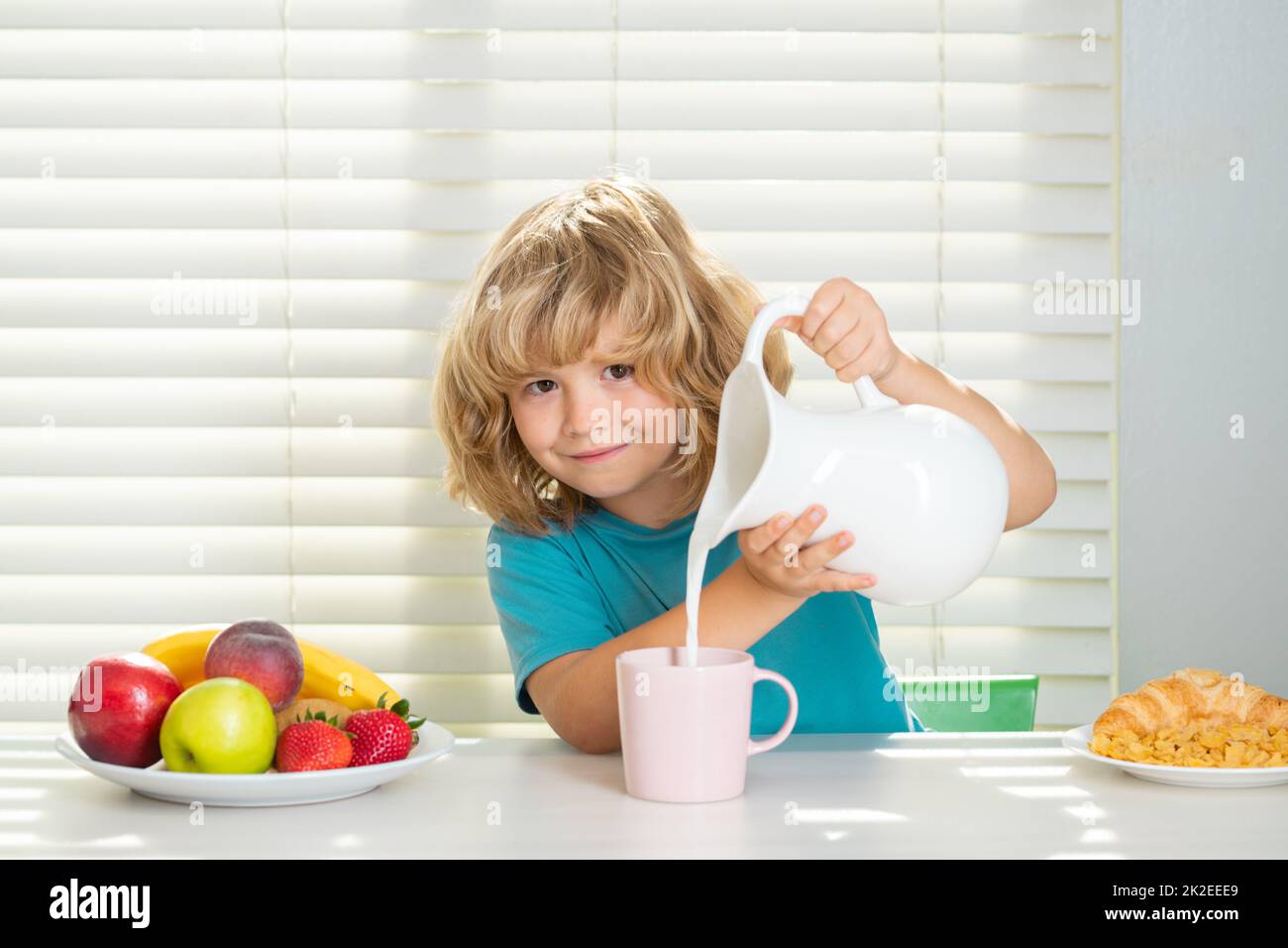 Kid boy pouring whole cows milk. Portrait of child eat fresh healthy food in kitchen at home. Kid boy eating breakfast before school. Stock Photo
