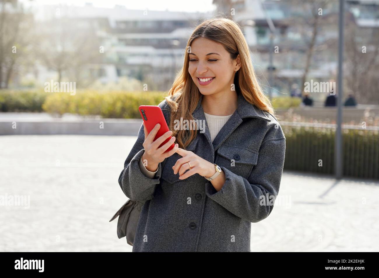 Medium shot of young smiling woman using cellphone in the street on sunny day Stock Photo