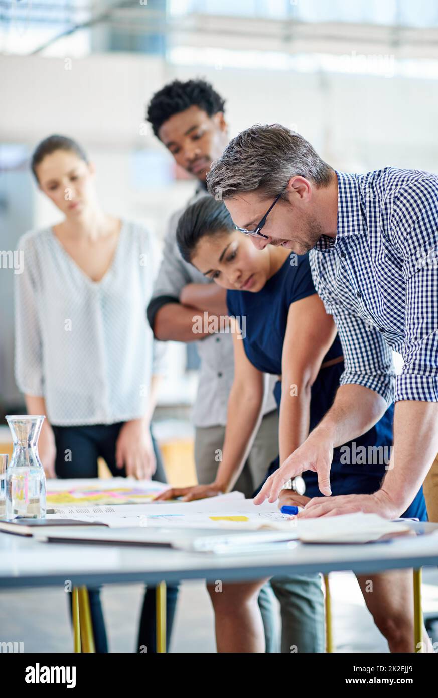 Leading the way with creative collaborations. Male and female managers planning at a table with younger staff members looking on. Stock Photo