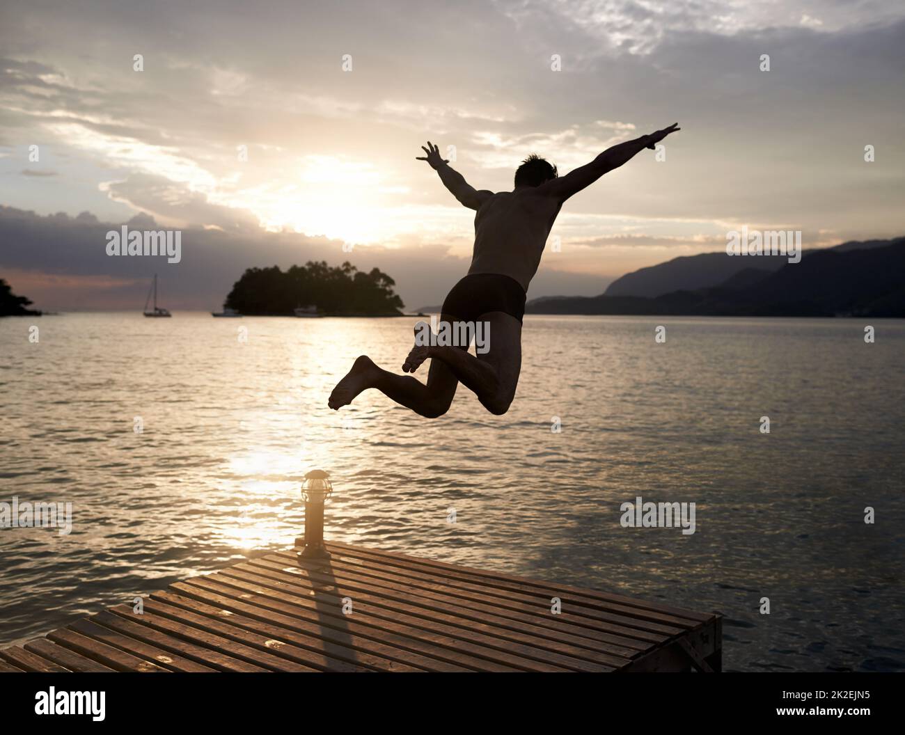 Spontaneity at sunset. Rear view shot of a young man diving off the jetty into a lake at sunset. Stock Photo