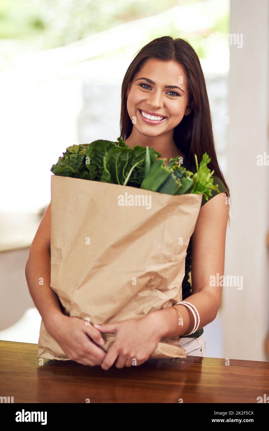 Eating well and feeling great. Portrait of an happy young woman holding a bag of groceries in her kitchen. Stock Photo