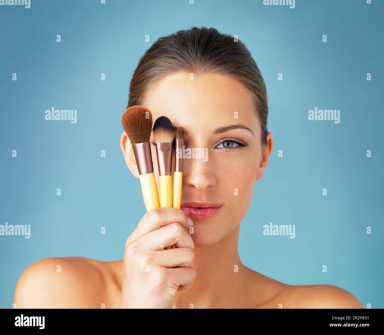 The secret to a flawless makeup routine. Studio portrait of a beautiful young woman holding makeup brushes in front of her eyes against a blue background. Stock Photo